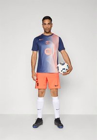 Camisola Nike Barcelona em azul com acentos laranja em degradê, combinada com calções laranja brilhante. O jogador segura uma bola de futebol com padrão.