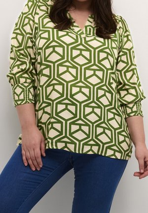 Kvinde iført grøn og cremefarvet bluse med geometrisk mønster og blå tætsiddende jeans mod en ensfarvet baggrund.