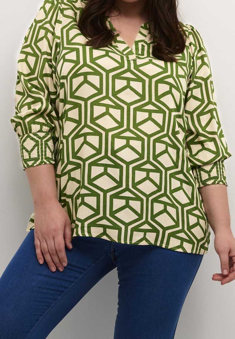 Femme portant un chemisier à manches longues à motifs géométriques verts et crème, associé à un jean bleu ajusté sur un fond uni.