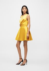 Γυναίκα με κίτρινο σατέν αμάνικο φόρεμα με πιέτες στη μέση και μαύρα ψηλοτάκουνα σανδάλια, που στέκεται μπροστά σε λευκό φόντο.