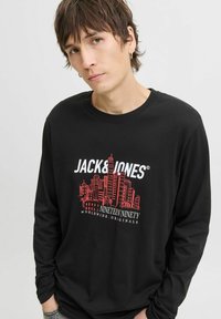 Joven con cabello corto castaño que lleva una camisa de manga larga negra con un skyline de la ciudad en rojo y el texto "JACK & JONES NINETEEN NINETY".
