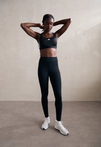 Fekete sportmelltartó fehér részletekkel és logóval, magas derekú fekete leggingssel párosítva. A modellen fehér sportcipők vannak. Simma, testhez álló textúra.