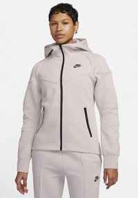 Lichtroze Nike zip-up hoodie met een aangesloten ontwerp, zwarte ritssluiting, twee zijzakken en een logo op de linkerborst; zachte stoffen textuur.