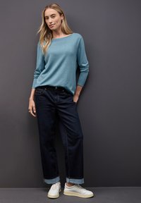 Maglione lavorato a maglia di colore azzurro chiaro con maniche lunghe, abbinato a jeans a gamba larga di colore blu scuro e sneakers bianche. I jeans hanno un orlo arrotolato.