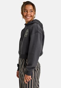Sudadera corta de color gris oscuro con capucha, que presenta un diseño de logo en el frente y puños acanalados, combinada con pantalones de tiro alto a rayas verticales.