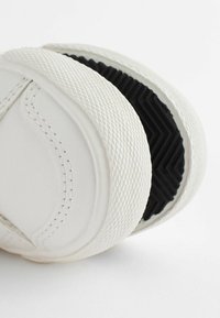 Hvide sneakers med en tekstureret overflade, gummisål og et sort zigzag mønster på bunden. Har syede detaljer og glatte kanter.
