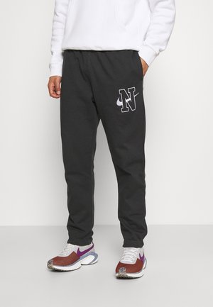 Mężczyzna ubrany w czarne spodnie dresowe z białym „N” i logo Nike, białą bluzę i białe-fioletowo-brązowe buty Nike, stojący przy białej ścianie.