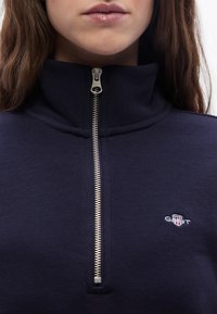 Κοντινό πλάνο ατόμου που φοράει μπλε σκούρο φούτερ με φερμουάρ και γιακά, με μικρό λογότυπο GANT στο στήθος.