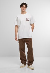 Biely bavlnený tričko s malou červenou a čiernou grafikou na hrudi, spárované s hnedými pohodlnými nohavicami a béžovými teniskami.