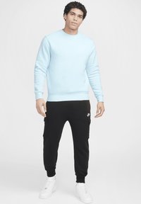 Sudadera azul claro con puños acanalados, combinada con pantalones cargo negros que tienen bolsillos laterales y un logo blanco, completada con zapatillas blancas.