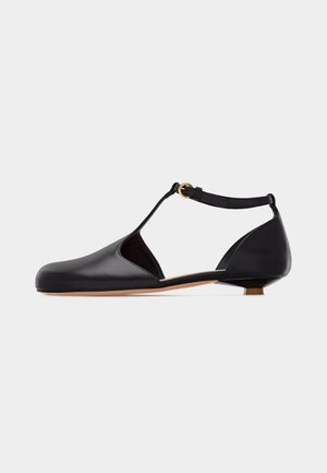 Black leather low-heeled T-strap shoe with rounded toe, cut-out sides, ankle strap, and gold-tone buckle on light gray background.