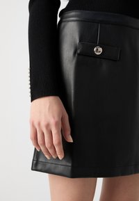 Jupe en cuir noir au design épuré. Elle dispose d'une poche avant avec un bouton-pression et de détails de couture visibles. Surface texturée.