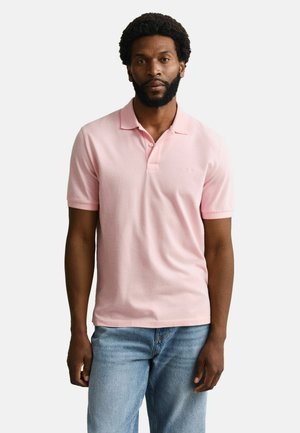 Mann mit Bart, der ein hellrosa kurzärmeliges Poloshirt und blaue Jeans trägt, vor einem schlichten weißen Hintergrund stehend.