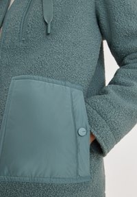 Polarowa kurtka w stonowanym odcieniu teal z teksturowanym korpusem, gładką kieszenią z guzikiem oraz zamkiem błyskawicznym przy szyi.