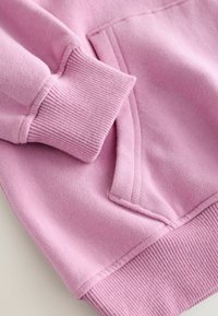 Närbild av en rosa sweatshirtärm och ribbad mudd bredvid en framficka med synlig sömnad och mjuk tygtextur.
