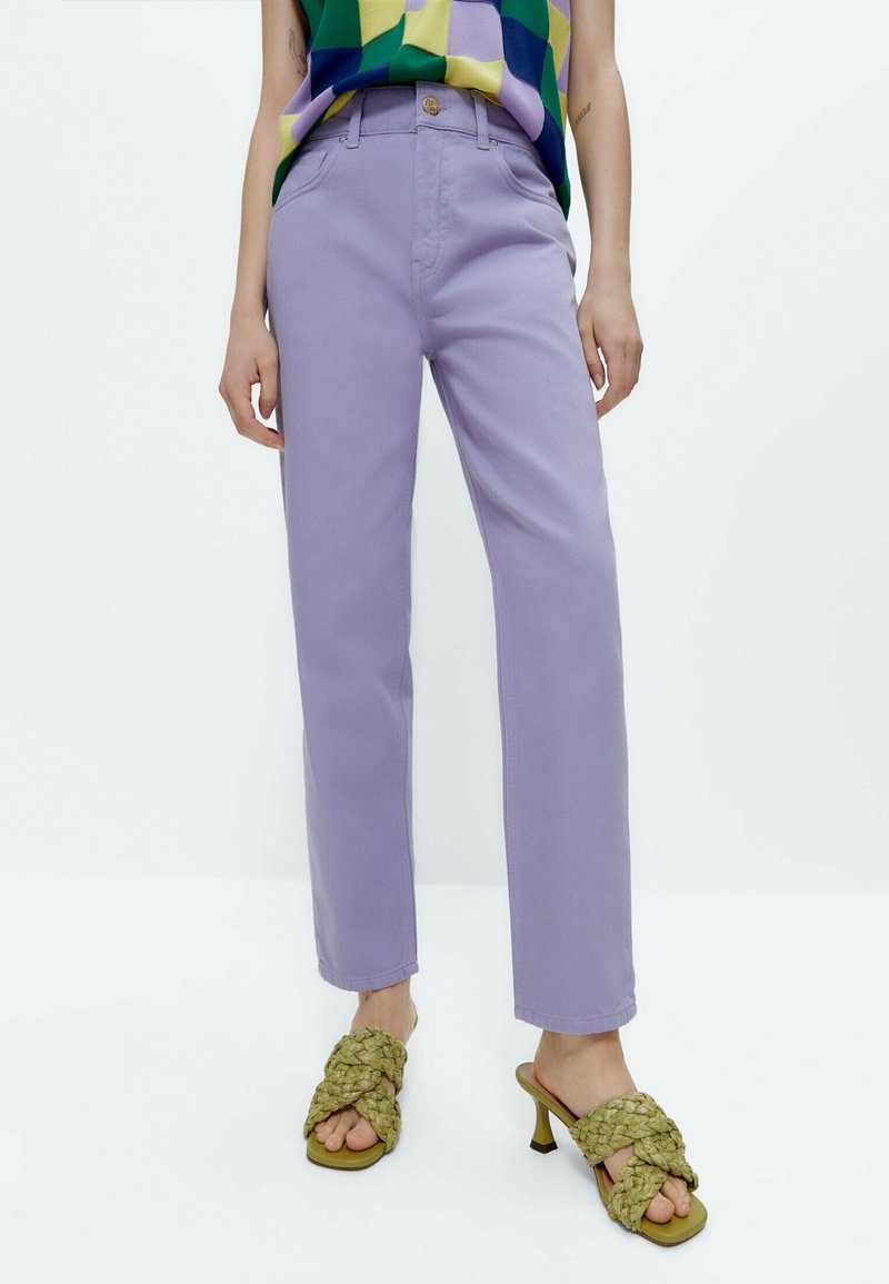 Pantalon en coton lavande à coupe droite, poches avant et bouton doré. Associé à des sandales à talons vertes tressées.