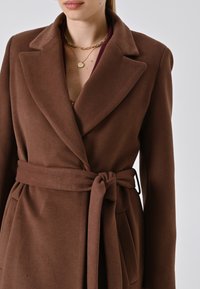 Donna che indossa un cappotto marrone con cintura e ampi rever, collane d'oro a strati e una maglia bordeaux sottostante.