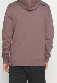 Sudadera con capucha en marrón apagado, hecha de tela suave. Presenta una capucha trasera, puños acanalados y un pequeño logo negro en el hombro.