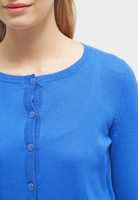 Blå stickad cardigan med rund halsringning, med en framknäppning och texturerad ribbning längs kanterna. Slät tygstruktur.