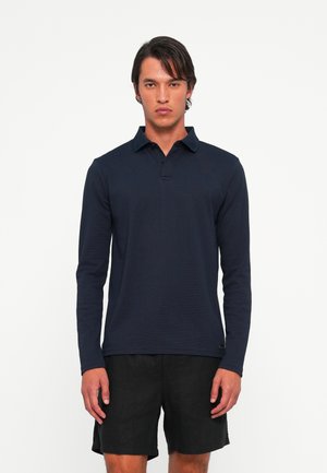 Man wearing a navy long-sleeve polo shirt and black shorts, standing against a plain white background, facing forward with hands relaxed.