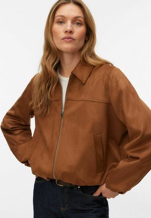 Mujer con cabello largo y rubio, que lleva una chaqueta marrón con cremallera y vaqueros oscuros, de pie con las manos en las caderas contra un fondo liso.