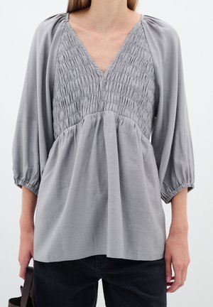 Kobieta ubrana w jasno szarą marszczoną bluzkę z dekoltem w serek i rękawami balonowymi oraz czarne spodnie, trzymająca w lewej ręce ciemny pasek do torby.