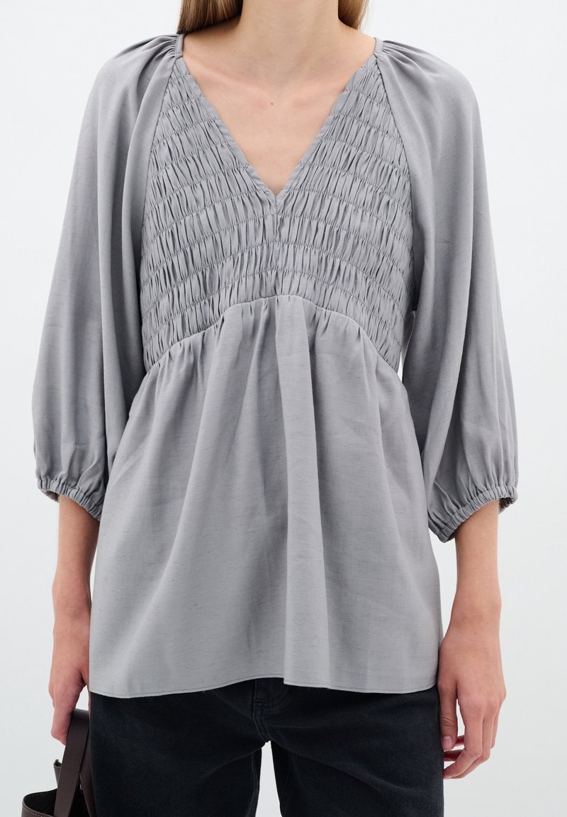 Femme portant un chemisier gris clair smocké à col en V avec des manches ballon et un pantalon noir, tenant une sangle de sac foncée dans la main gauche.