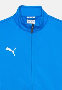 Plava sportska jakna na patent s visokim ovratnikom i bijelim Puma logotipom na lijevoj strani prsa.