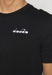 Černé bavlněné tričko s kulatým výstřihem a krátkými rukávy, se bílým vyšívaným logem na levé části hrudníku.