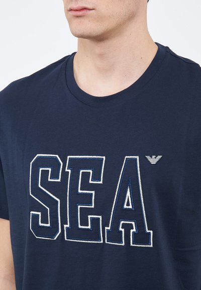 Giovane uomo che indossa una T-shirt blu navy con grandi lettere ricamate "SEA" e un piccolo logo di un'aquila sopra la lettera "A" sul petto.