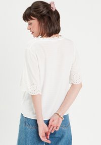 White short-sleeve top with subtle dot texture, ruffled neckline, and lace-patterned accents at sleeves. Worn with blue denim jeans.
