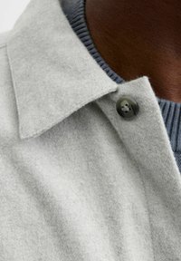 Abrigo de lana gris claro con un amplio cuello y un escote acanalado gris oscuro. Tejido texturizado con un acabado suave. Detalle de botones visible.