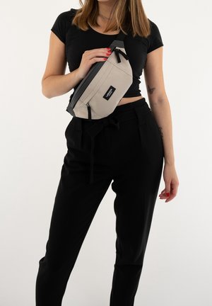 Kvinde iført sort crop top og bukser, der holder en beige og sort crossbody taske med mærket "LARKSON" tværs over brystet.