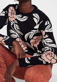 Schwarzer Pullover mit floralem Design in Rosa und Weiß, gerippter Rundhalsausschnitt und Bündchen, kombiniert mit rostfarbenen Hosen. Weicher Stoff, Strickmuster.