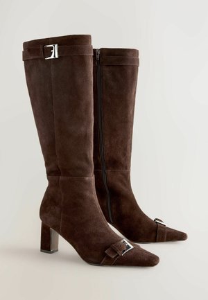 Ein Paar kniehohe Stiefel aus dunkelbraunem Wildleder mit spitzen Zehen, mittelhohen Absätzen, seitlichen Reißverschlüssen und silbernen Schnallenakzenten an den oberen und vorderen Riemen.