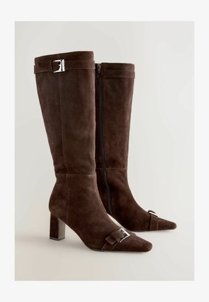 Ein Paar kniehohe Stiefel aus dunkelbraunem Wildleder mit spitzen Zehen, mittelhohen Absätzen, seitlichen Reißverschlüssen und silbernen Schnallenakzenten an den oberen und vorderen Riemen.