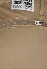 Beige denim tyg med en diagonal väv, en silver dragkedja ficka och en vit etikett med "G-Star Originals RAW Denim" tryckt i svart.