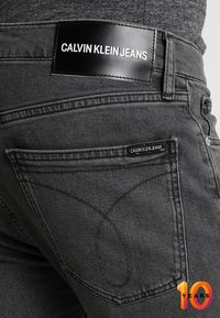 Vaqueros Calvin Klein en denim gris oscuro, con una etiqueta negra con texto blanco, costuras detalladas y un diseño de bolsillo trasero.