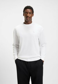White long-sleeve shirt with a crew neck and a small logo detail on the chest, paired with black trousers. Smooth fabric texture.