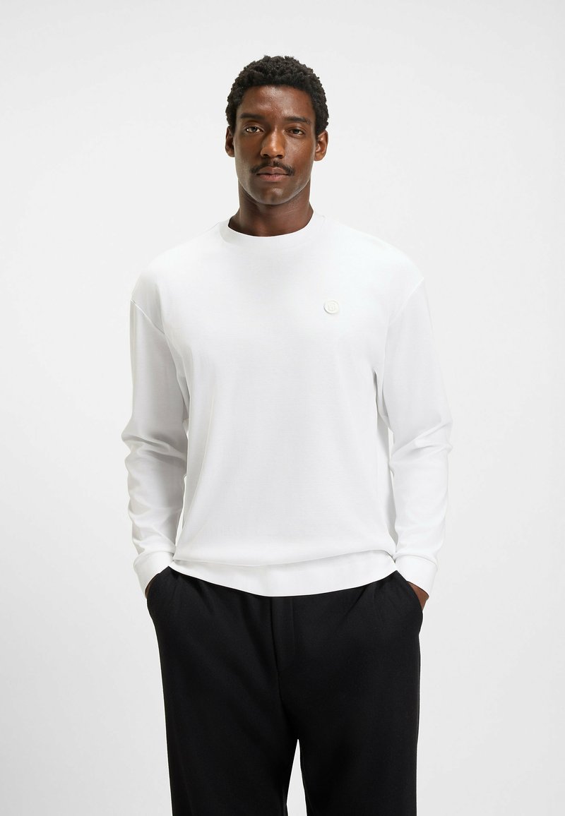 White long-sleeve shirt with a crew neck and a small logo detail on the chest, paired with black trousers. Smooth fabric texture.