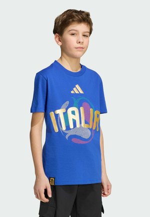 Garçon debout portant un t-shirt bleu « Italia » avec un design coloré et un short cargo noir, sur un fond clair uni.