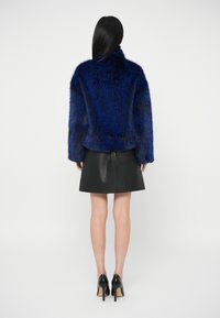 Γυναίκα με μακριά μαύρα μαλλιά, φορώντας μια μπλε γούνινη ζακέτα, δερμάτινη φούστα μαύρου χρώματος και μαύρες γόβες, στέκεται με την πλάτη γυρισμένη.