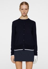 Merenkookedista valmistettu pehmeä neulejakku, jossa on pyöreä pääntie, nappikiinnitys, sekä kontrastia tehostava valkoinen viimeistely helman ja hihansuiden kohdalla.