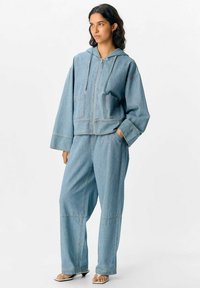 Giacca di jeans azzurro chiaro con cappuccio e zip frontale, abbinata a pantaloni larghi coordinati con tasche laterali e cuciture a vista.