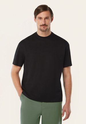 Mann mit Schnurrbart und kurzen Haaren, der ein schlichtes schwarzes T-Shirt und grüne Hosen trägt, mit einer Hand in der Tasche vor hellem Hintergrund.