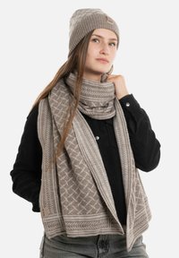 Beige gebreide muts en sjaal set met geometrische patronen, zachte textuur en een labeltag, gedragen over een zwart geribbeld sweater.