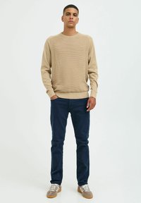 Pull en tricot beige avec texture côtelée, col rond et manches longues, associé à un pantalon bleu marine et des baskets gris-brun.