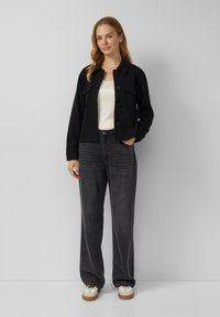 Vrouw in een zwarte knoopjasje, witte top, losse donkergrijze jeans en witte sneakers, staande tegen een effen lichtgrijze achtergrond.