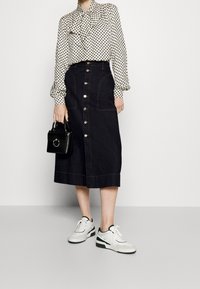 Kvinde iført en mønstret bluse, en knaplukket sort midi-nederdel, hvide sneakers og holder en lille sort håndtaske med en metalring.