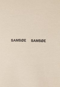 Beige stof met de tekst "SAMFØE SAMFØE" in vet zwarte letters, met een gladde, geribbelde textuur en een minimalistisch ontwerp.
